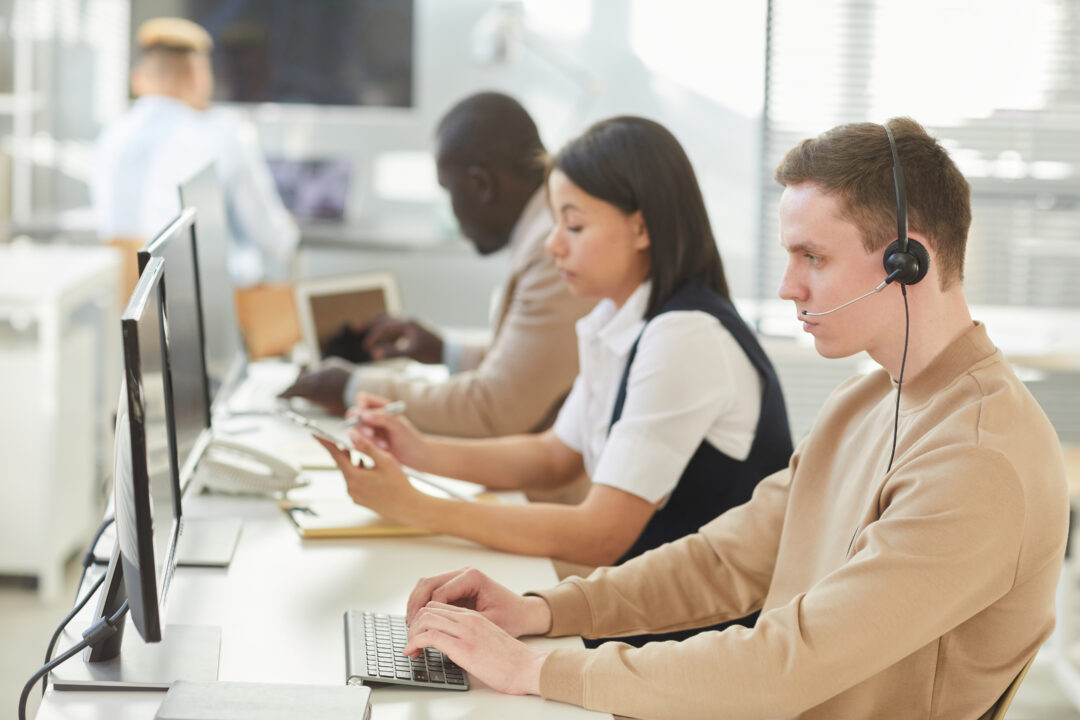 Young people wearing headsets sitting in row while working in call center